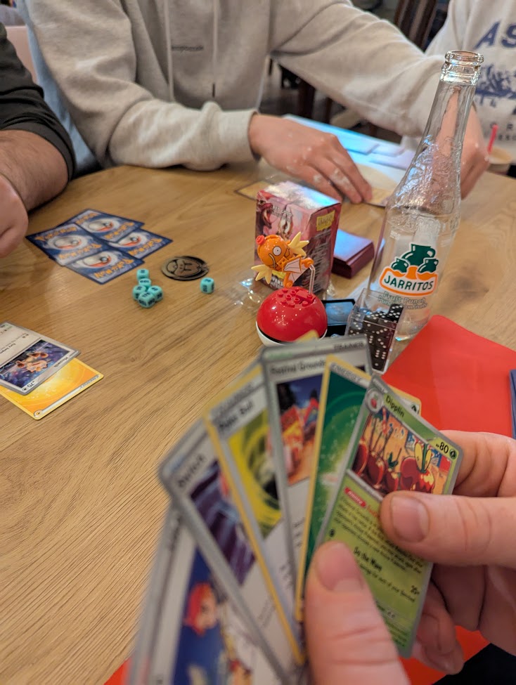 An image of a tournament table. There's a hand of cards in the foreground, other players in the background. In focus, near the middle, is a Magicarp figurine.